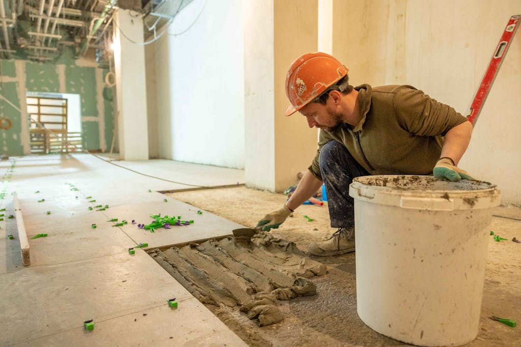 A construction worker lays tiles in an indoor renovation project, wearing safety gear.